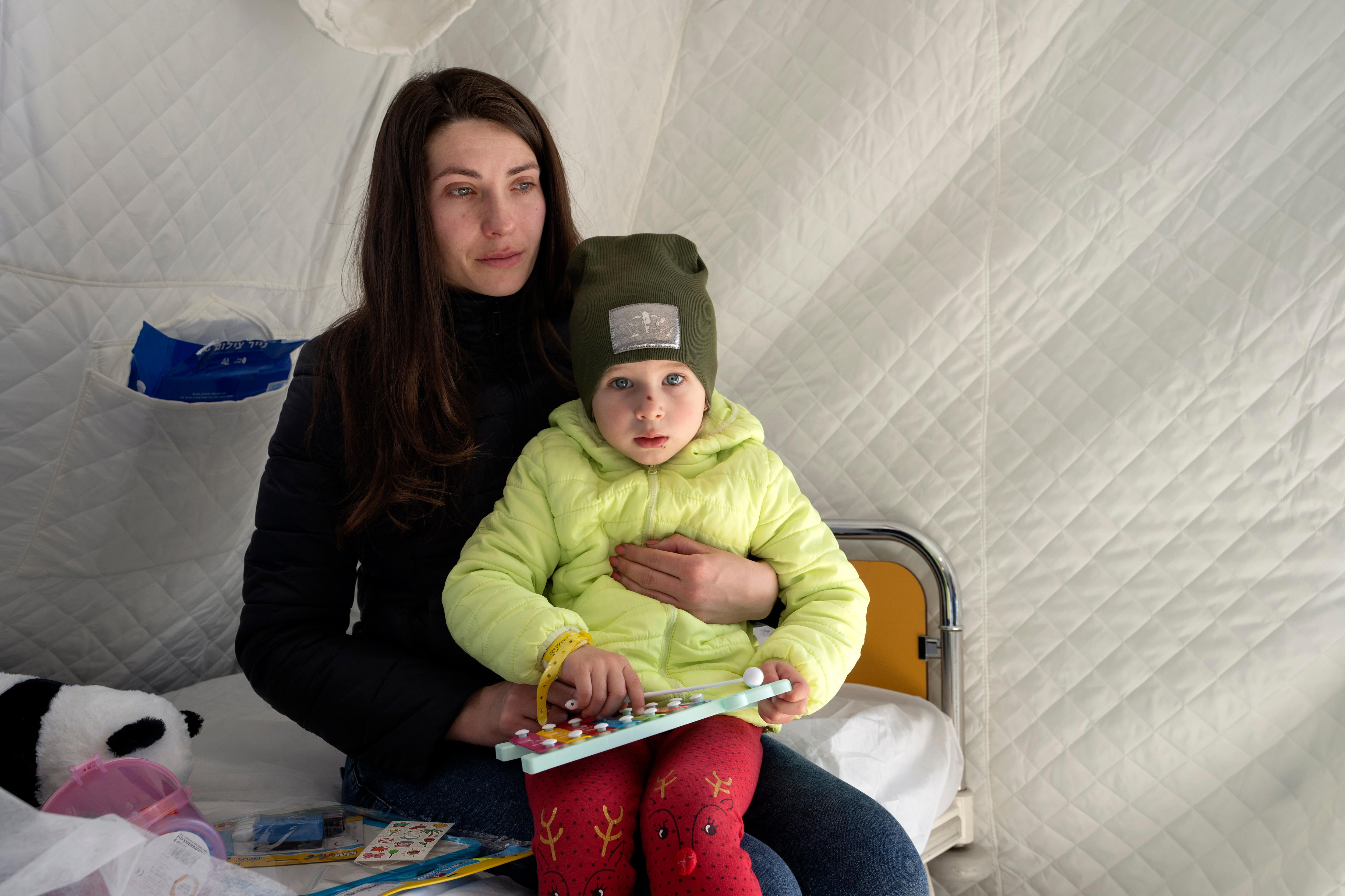 A three-year-old girl staring at the camera is held by her mother who looks away, sadly.
