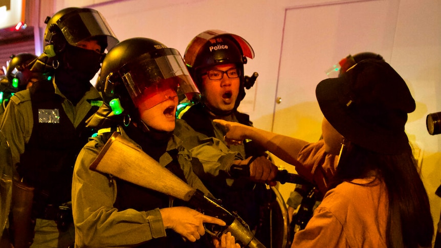 A close up photo shows a young woman in a bucket chat pointing at three police officers in riot gear carrying rifles.