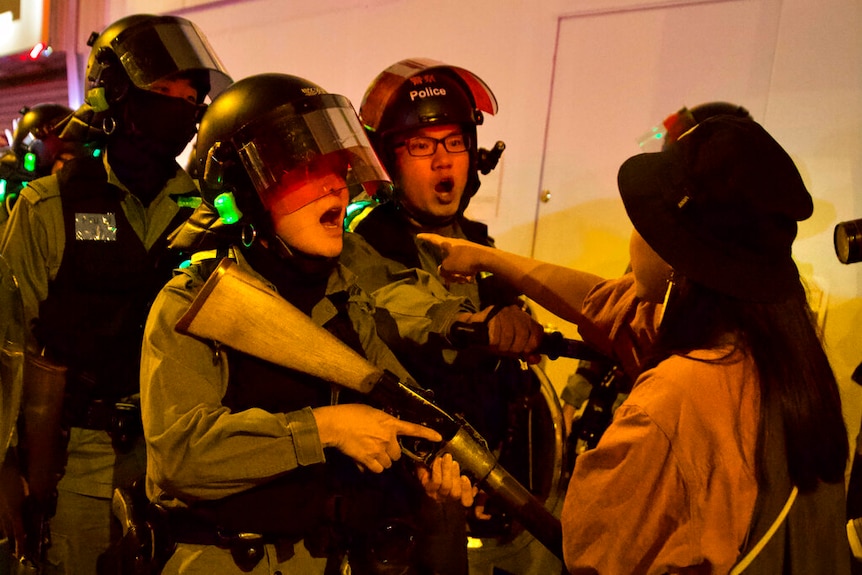 A close up photo shows a young woman in a bucket chat pointing at three police officers in riot gear carrying rifles.