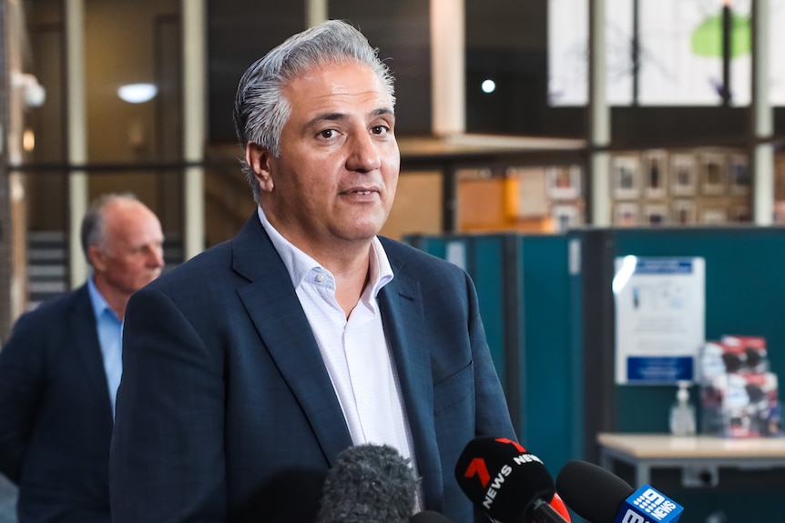 A man in a suit speaking with several microphones in front of him