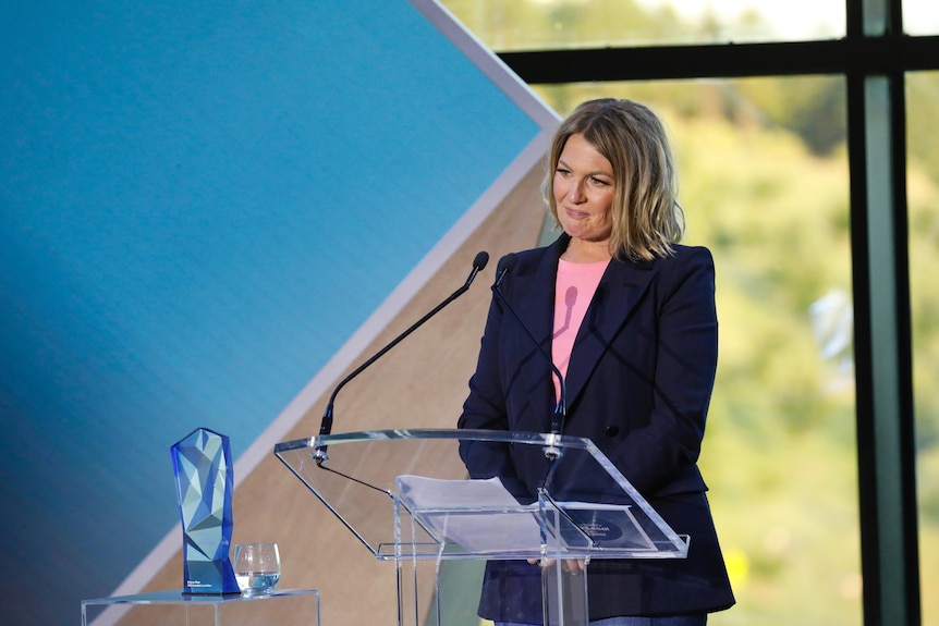 A woman standing at a lectern, smiles looking at the crowd. 