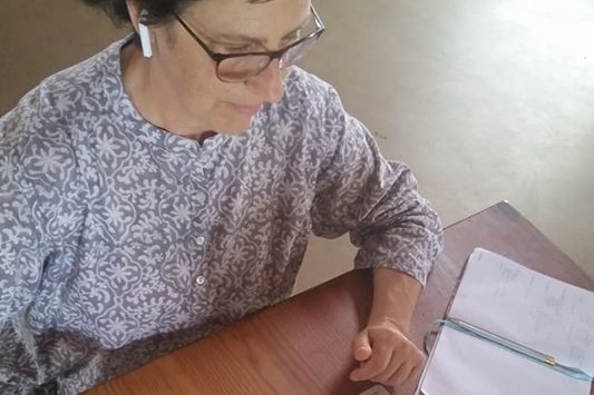 An older woman sits at a table with a laptop and cup of tea.