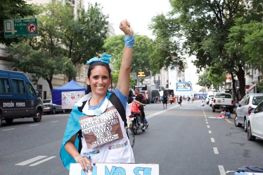 A woman wearing a blue ribbon in her hair and holding protests signs holds her fist in the air