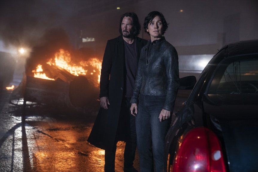 A 57-year-old man with long dark hair and beard stands next a 52-year-old woman, both in dark outfits, in front of a burning car