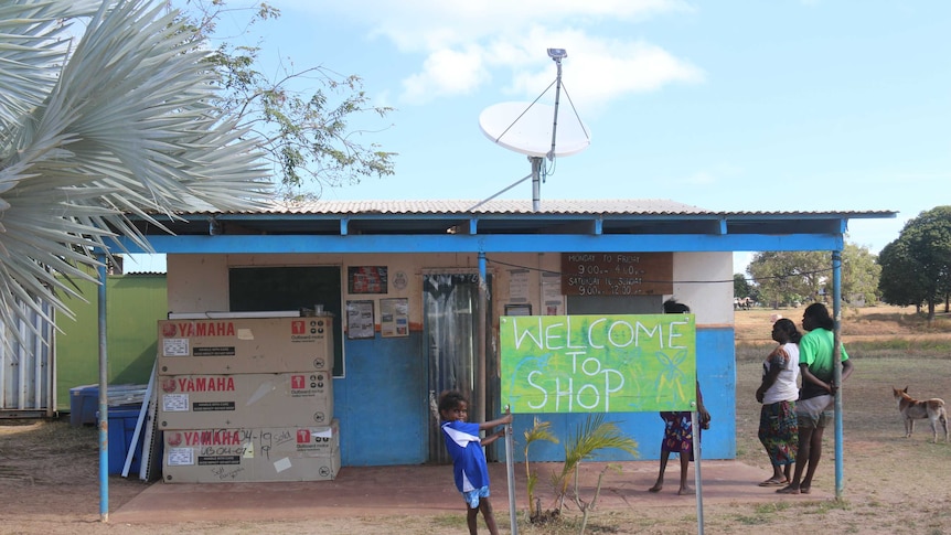 A shop front in Baniyala Indigenous community in Arnhem land with a green sign and a small child swinging on the porch column.