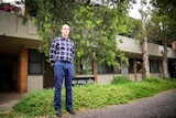 A balding middle-aged man in checked shirt and jeans stands with a serious expression in front on grey brick housing block