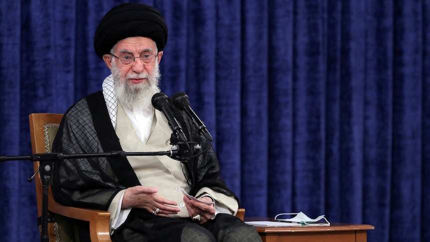 An old man in religious garb and head dress sits in a chair and speaks into a mic.