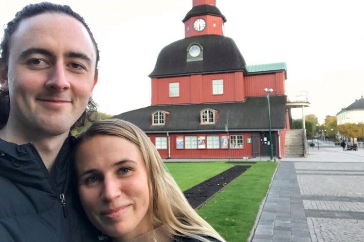 Ethan Brooker and his partner stare at the camera and embrace in a selfie taken in front of a big old red building.