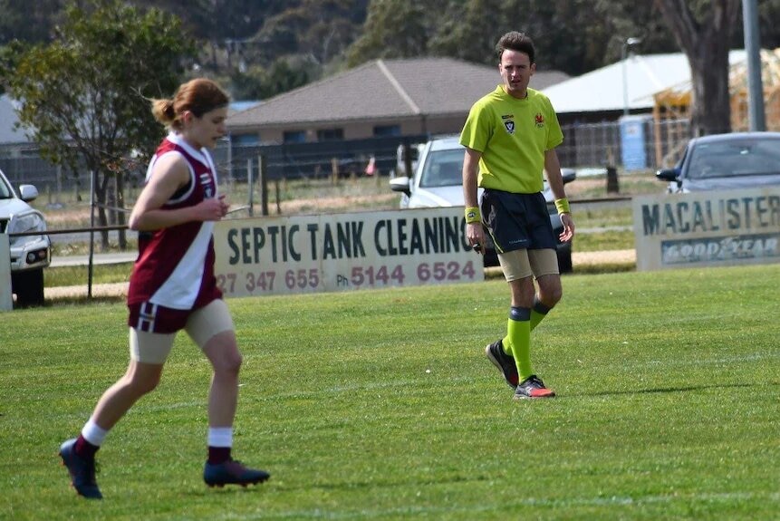 An umpire watches a player jog back to position in a game of junior women's football.