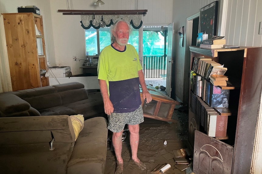 Rob Edgeworth stands in Maryborough home after flood surrounded by muddy furniture