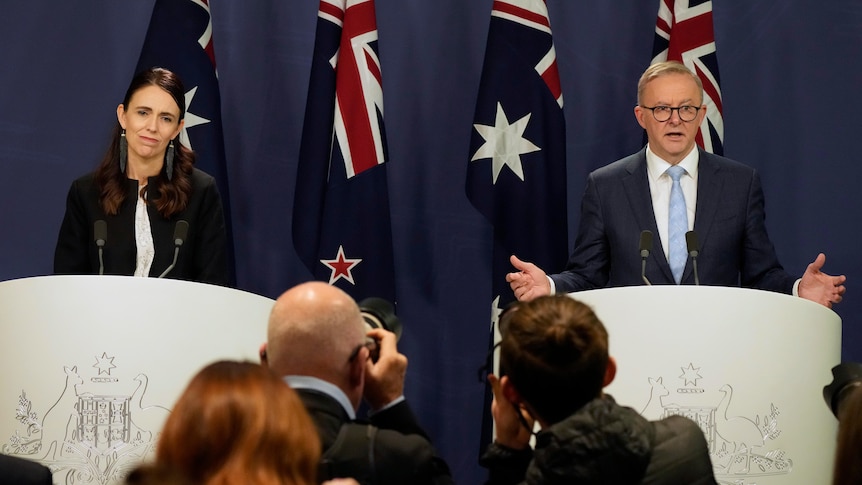 Two prime ministers stand at lecturns with their respective country's flags behind them. 