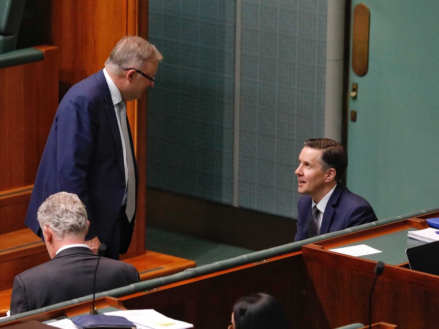 A man with grey hair and glasses in a suit and tie looks at another man in a suit and tie who's sitting on a bench in parliament