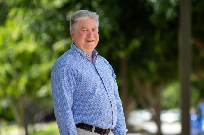 A man with grey hair smiles. He is wearing a blue cheque shirt and sand coloured pants. There is foliage in the background.