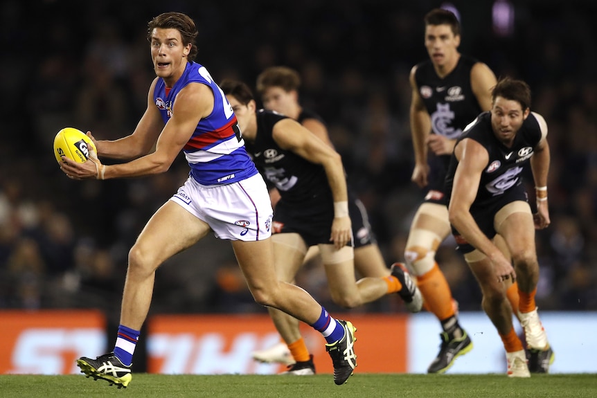 An AFL player holds the ball and looks upfield as he runs with defenders struggling to keep up behind him.