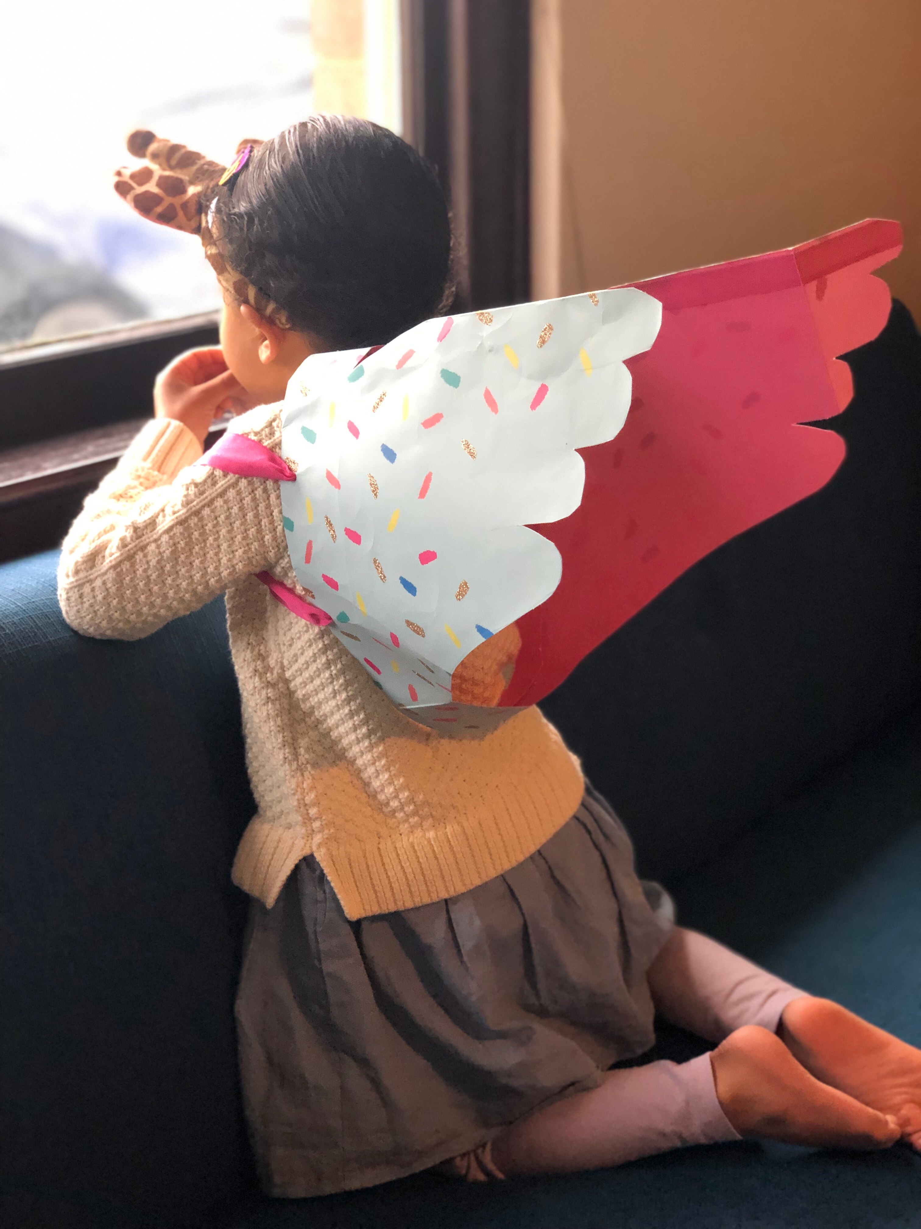 A girl looks out the window and wears unicorn wings made out of a pink gift bag.