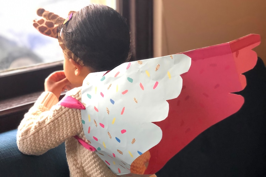A girl looks out the window and wears unicorn wings made out of a pink gift bag.