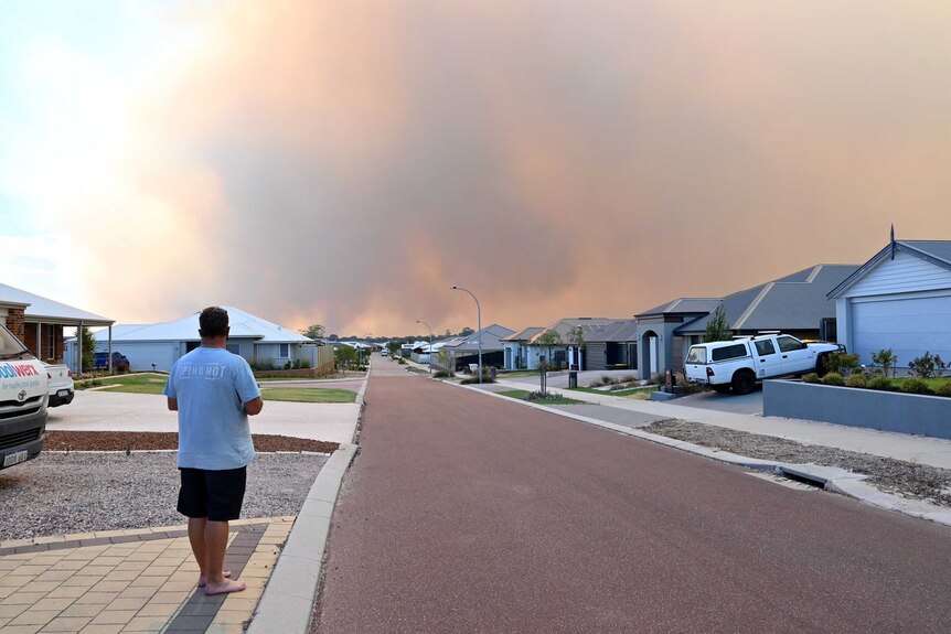 A man stands at the end of his driveway with heavy smoke in the sky.
