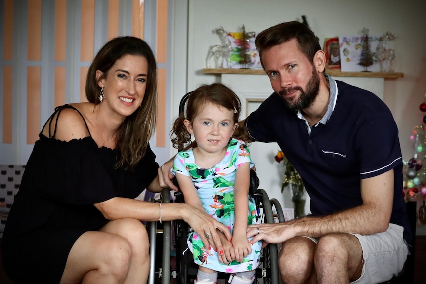 A young girl smiles to the camera. She is wearing a green dinosaur dress and uses a wheelchair. Her parents smile next to her.