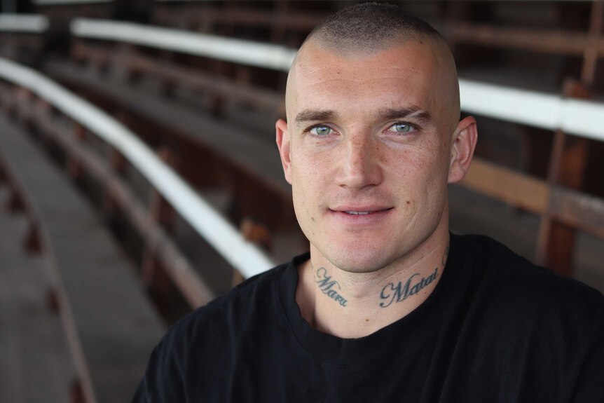 A portrait of a man with a throat tattoo and shaved head sitting on grandstand