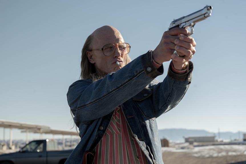 A film still of Ed Harris pointing a gun. He has a cigarette in his mouth, glasses, and is bald, but with long hair.