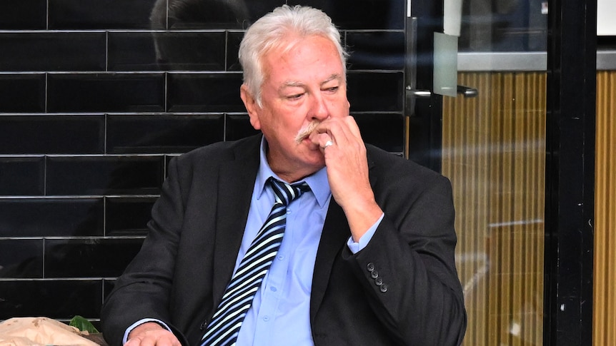 An older man wearing a dark suit chews his nails as he suits at a small cafe table