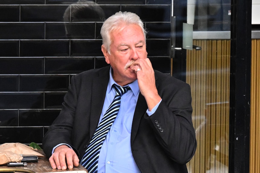 An older man wearing a dark suit chews his nails as he suits at a small cafe table