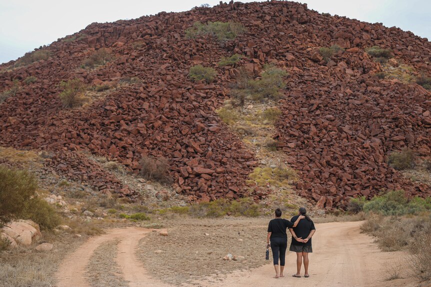 Two women stand facing away from the camera, looking at a large pile of red rocks with small green shrubs dotted throughout