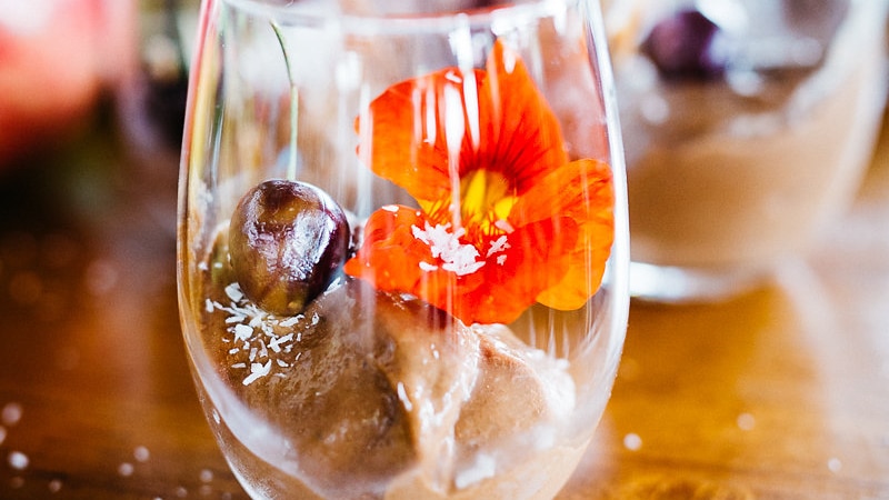 Chocolate mousse in a round glass on table with a cherry and also flower inside the glass.