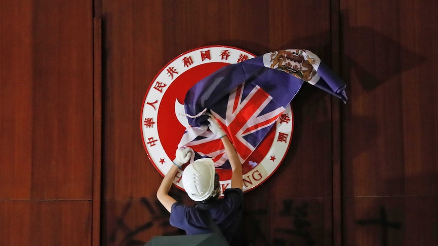 A protestor, flanked by two people with black umbrellas, covers the Chinese emblem of Hong Kong with the British Hong Kong flag.