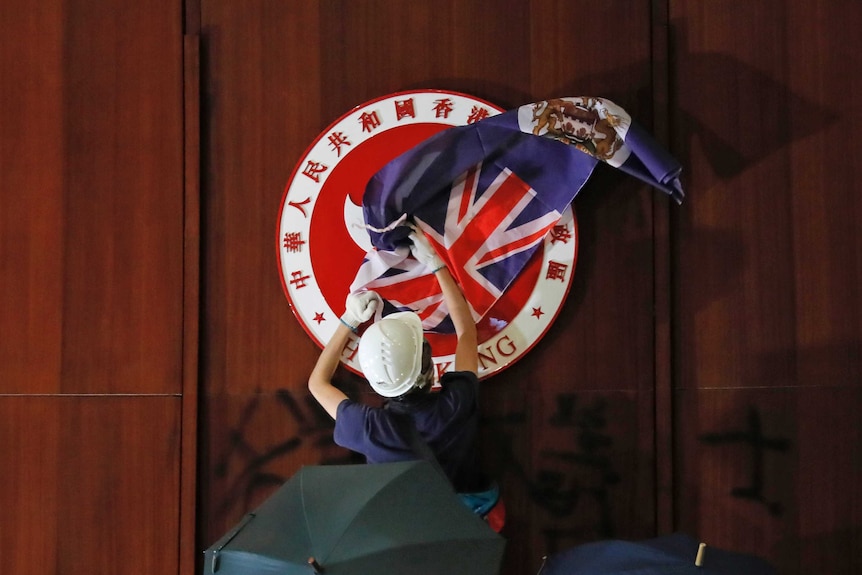 A protestor, flanked by two people with black umbrellas, covers the Chinese emblem of Hong Kong with the British Hong Kong flag.