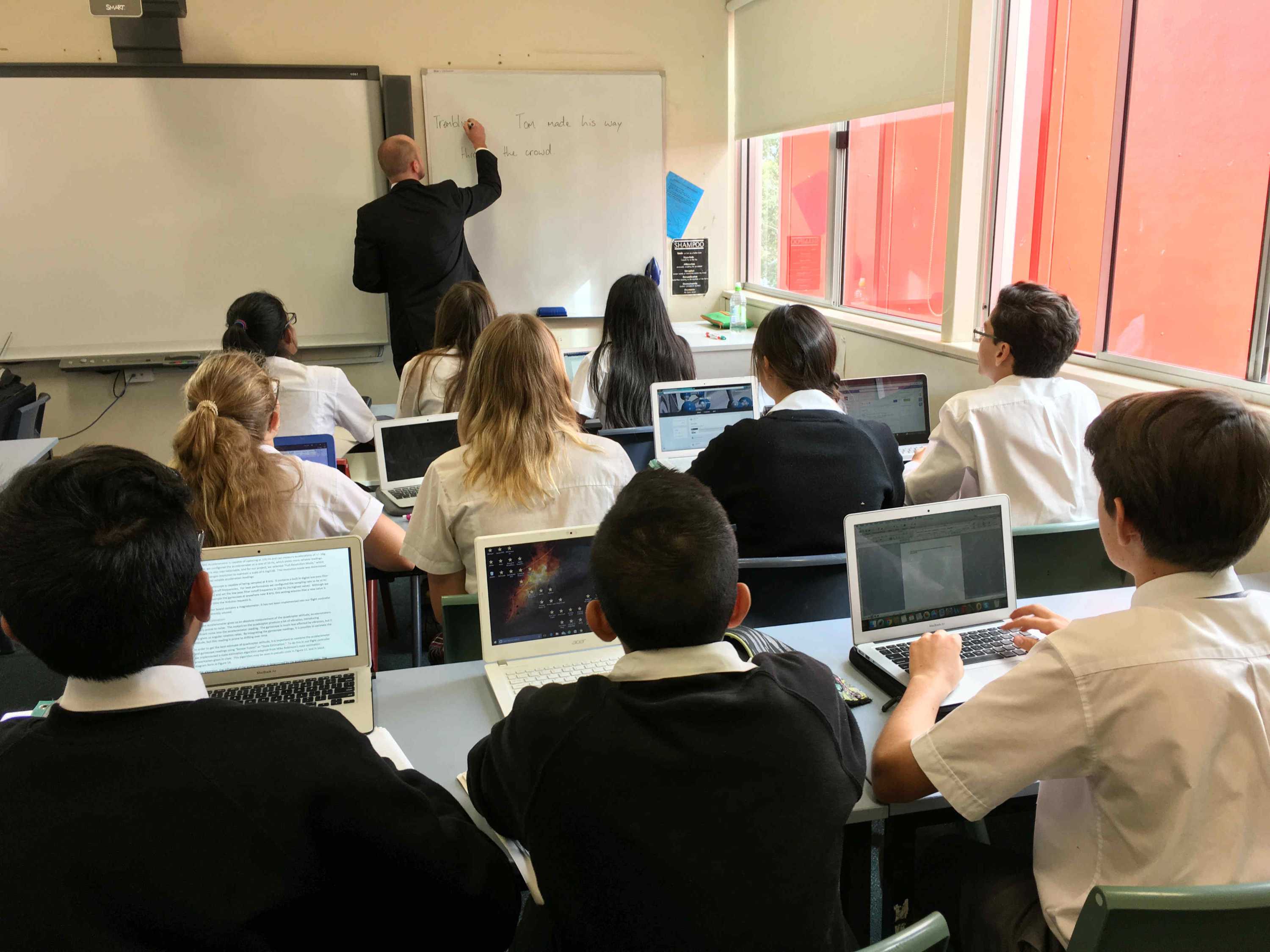 Students sitting at their desks watching a teacher write on a whiteboard