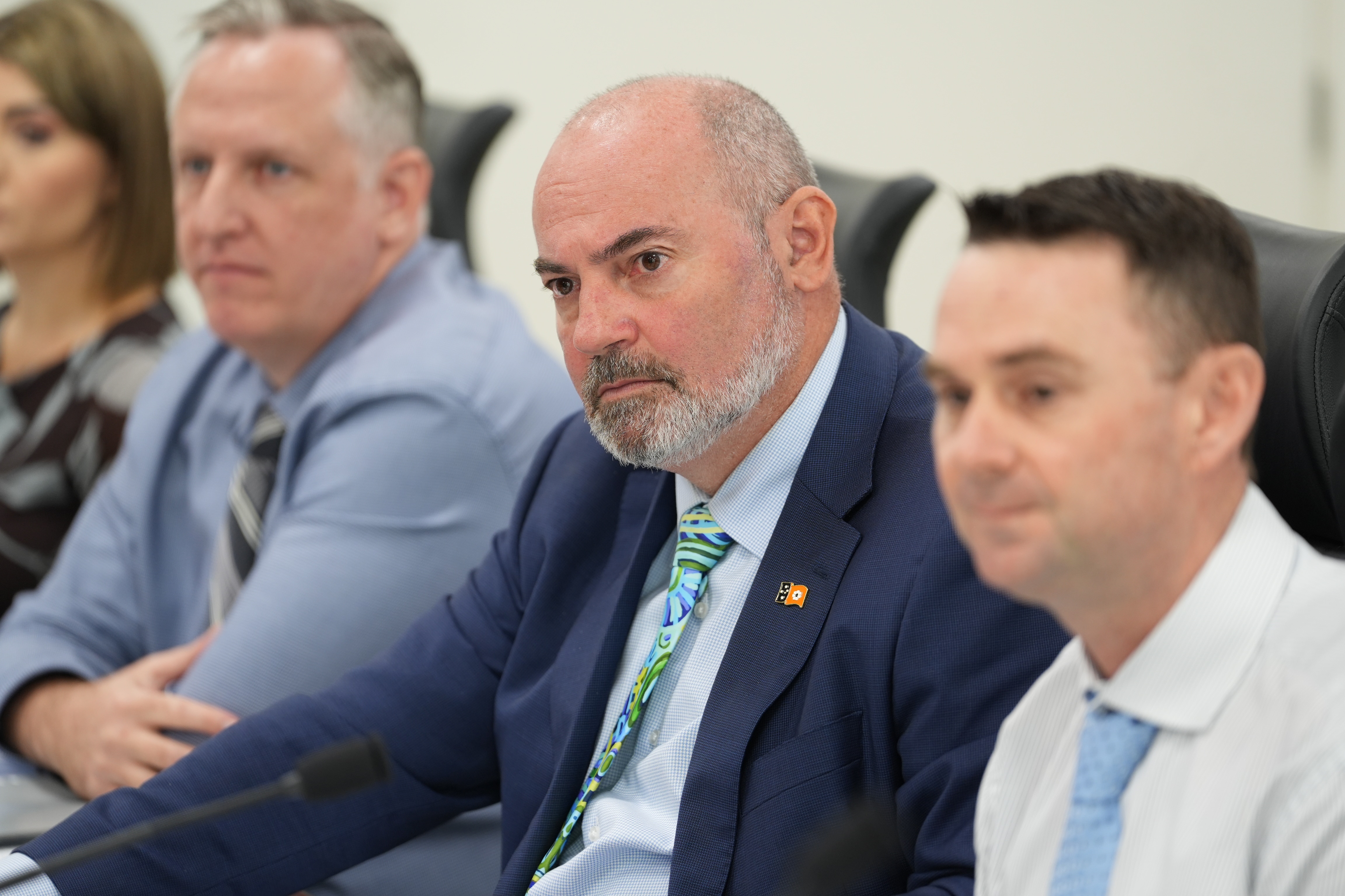 One man in focus sitting at table with out of focus other men in shirts and ties. Man has green / teal tie, serious expression.