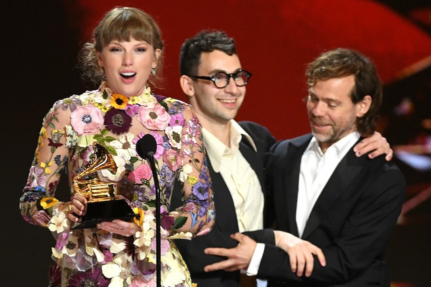Taylor Swift looks surprised on stage holding a Grammy award while Jack Antonoff and Aaron Dessner embrace behind her