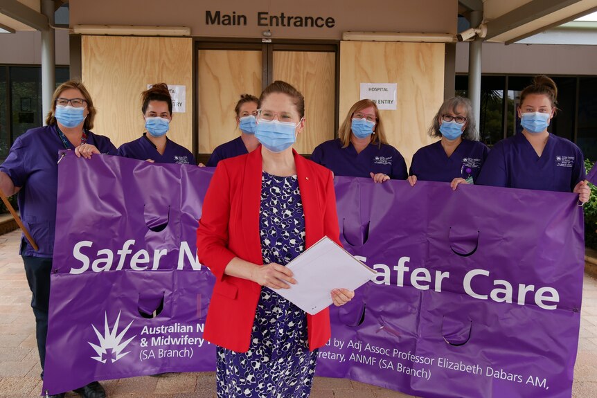 Nurses holding a purple sign reading 'Safer Nurses Safer Care' and a woman in a mask standing outside a damaged hospital.