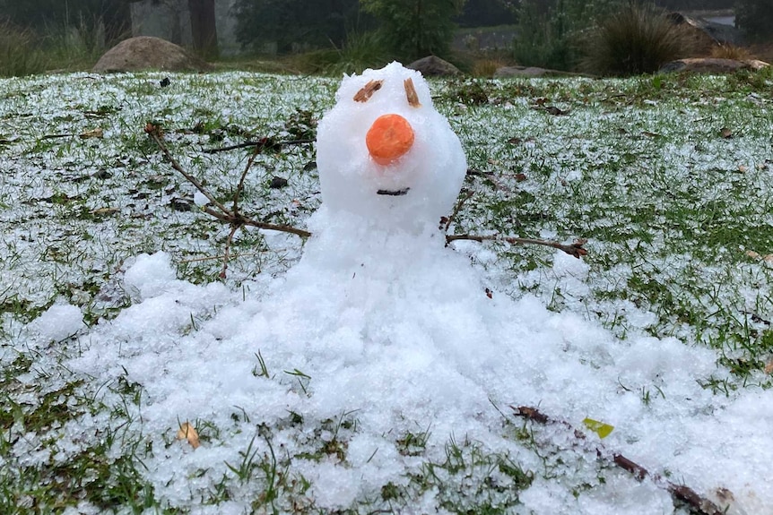 Tiny little snowman sitting on a slightly snow covered lawn where the grass can defiantly still be seen though.