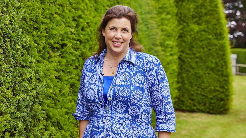 UK TV presenter Kirstie Allsopp, who has brown hair and wearing a blue dress, stands next to a row of hedges.