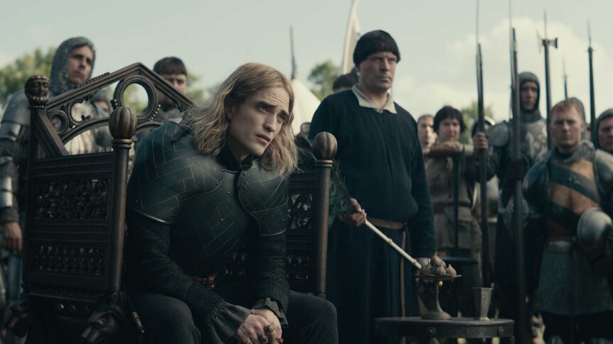 The actor Robert Pattinson dressed in full armour sitting on a throne surrounded by men in armor with spears