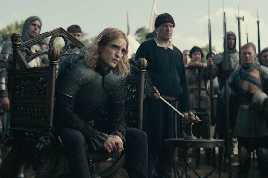 The actor Robert Pattinson dressed in full armour sitting on a throne surrounded by men in armor with spears