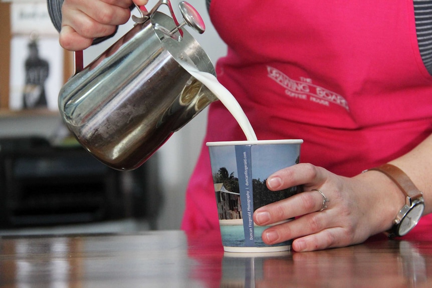 A mid shot of a girl (face not pictured) pouring hot milk from a steel jug into a takeaway cup.