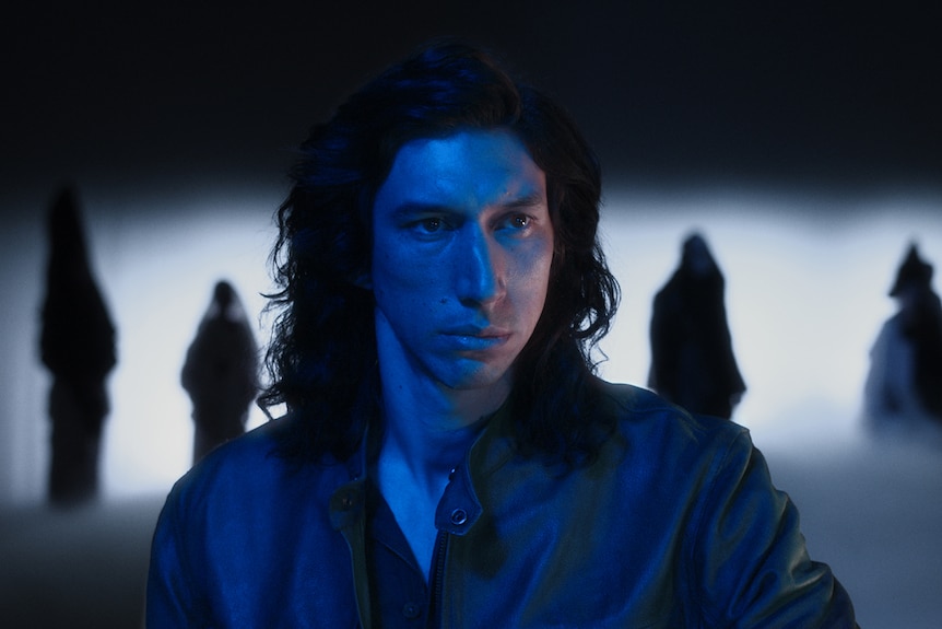 Adam Driver with shoulder-length dark hair is silhouetted by ghoulish figures. Blue light illuminates the concern on his face.