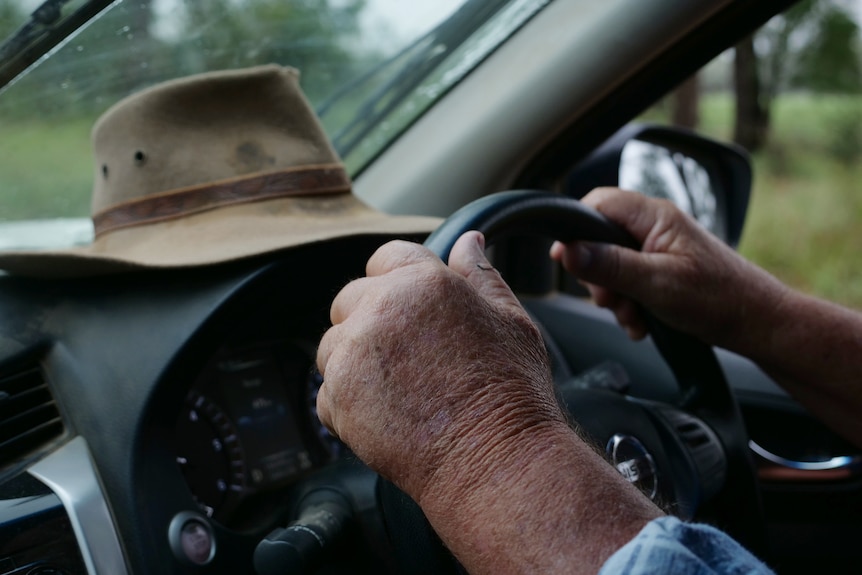 A man's weathered hands hold on to a steering wheel