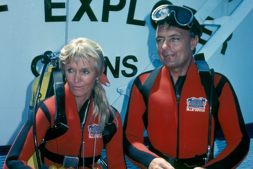 A man and a woman are seated while wearing red scub diving suits. The man has goggles on his head, the woman an air tank on back