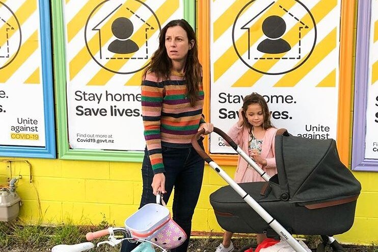 Anita Affleck pushes a pram and holds a child's bike in front of posters about coronavirus.