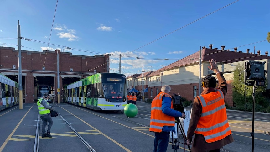 a tram in a depot approaching a large green ball being filmed by a crew in high viz vests