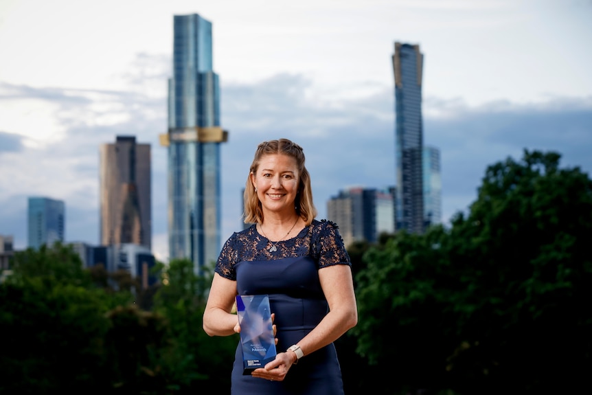 A woman holding a blue rectangular trophy smiles in front of skyscrapers.