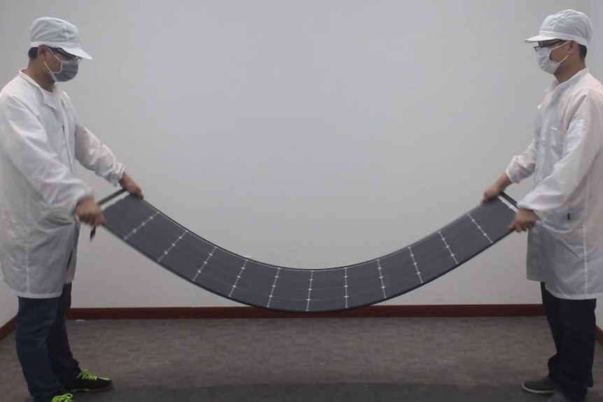 Two scientists, wearing masks and white lab coats, bending an experimental solar panel.