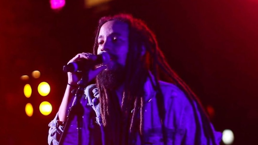 A Jamaican man with dreadlocks sings into a microphone onstage.