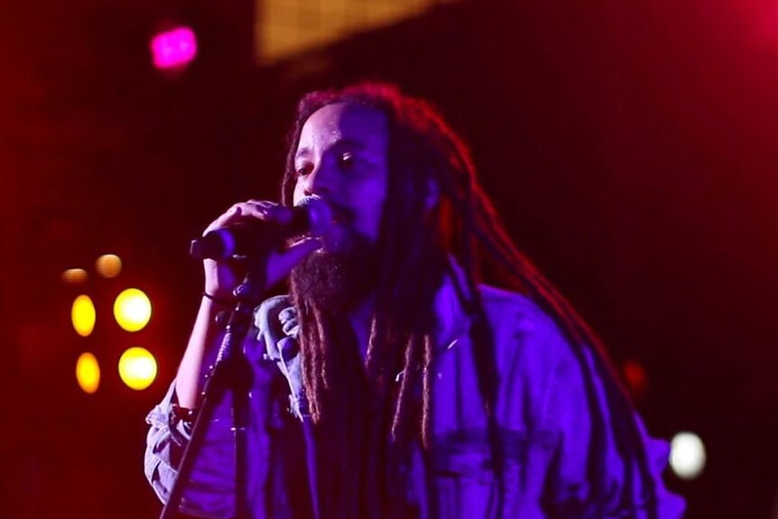 A Jamaican man with dreadlocks sings into a microphone onstage.