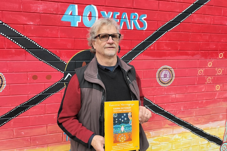 An indigenous man holding up a dictionary against an Aboriginal artwork.