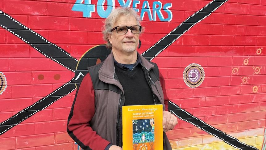 An indigenous man holding up a dictionary against an Aboriginal artwork.